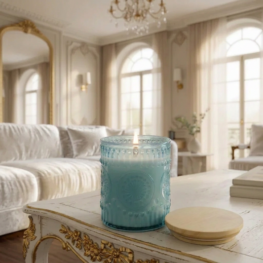 A light blue textured glass candle jar sits on a light wood table in a bright, modern living room with a blurred sofa and arched windows in the background.